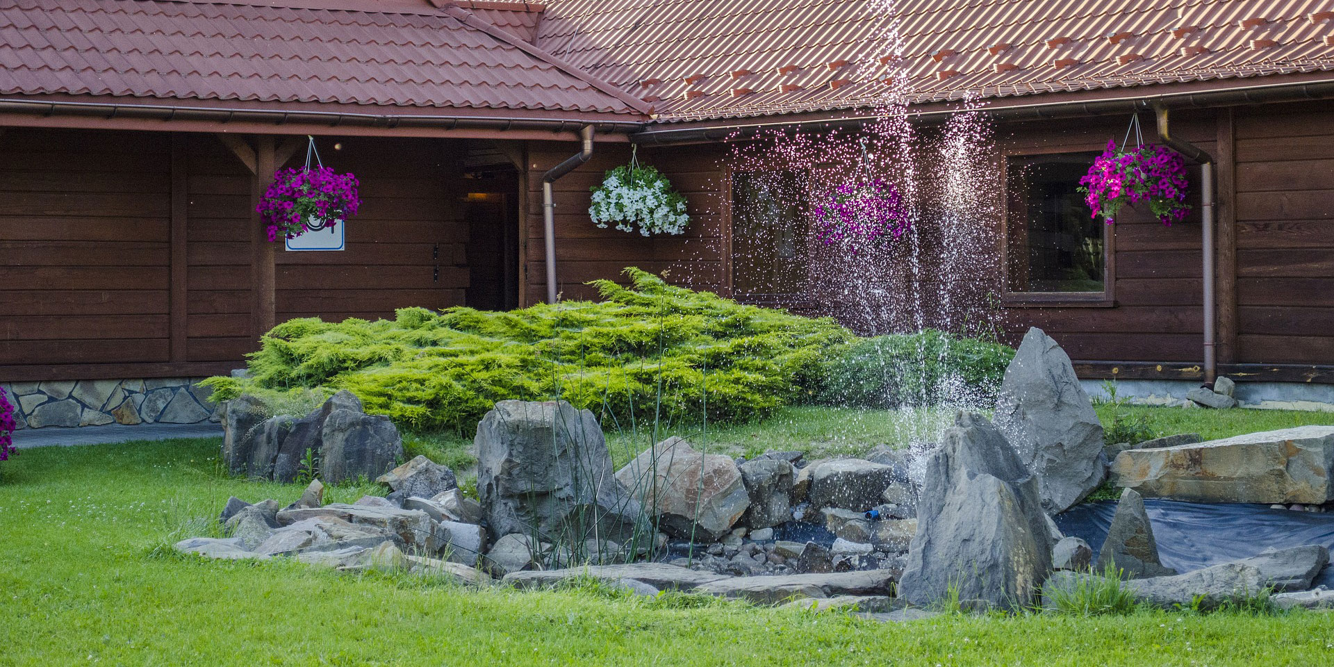 Fountain in rock surrounded pond with lawn and shrubs behind house